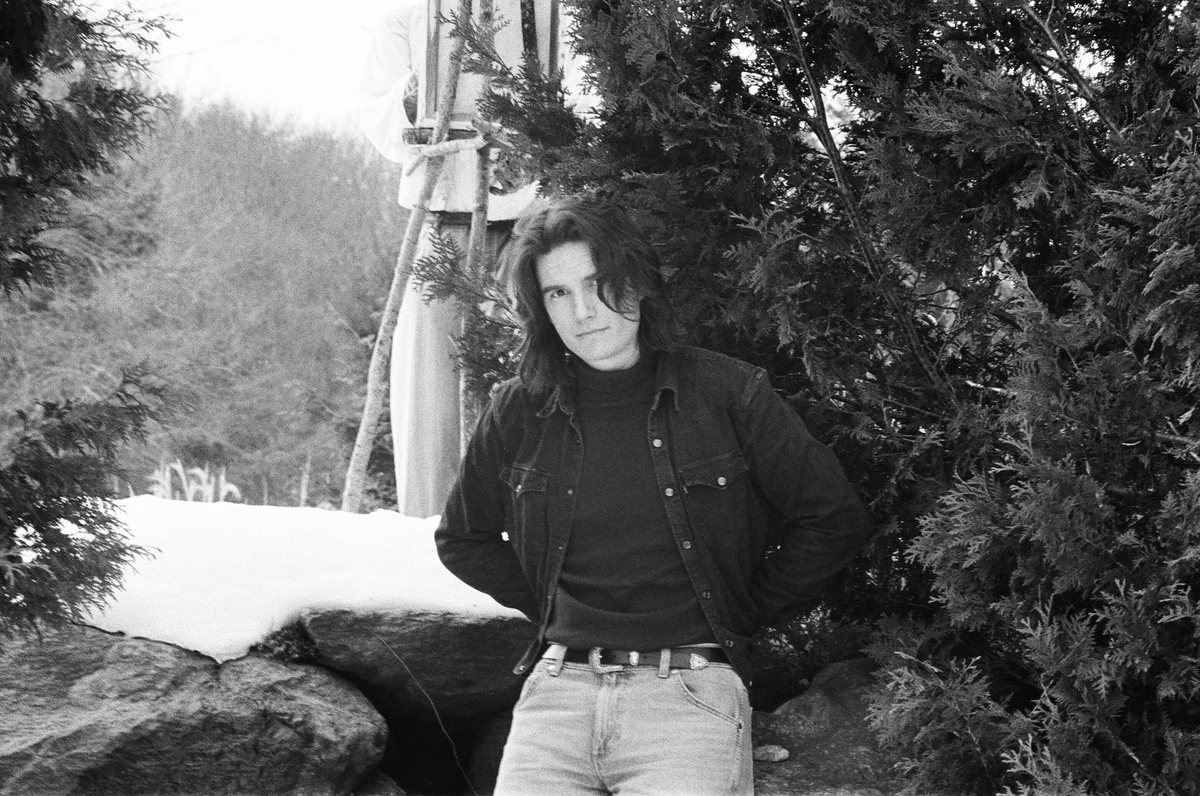 Greg Hall leaning against snowy rocks between evergreens, black and white